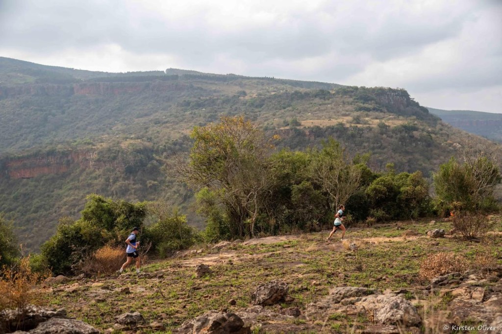 Runners on a trail below a rocky escarpment
